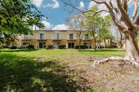 a view of a white house in front of a big yard with plants and large trees