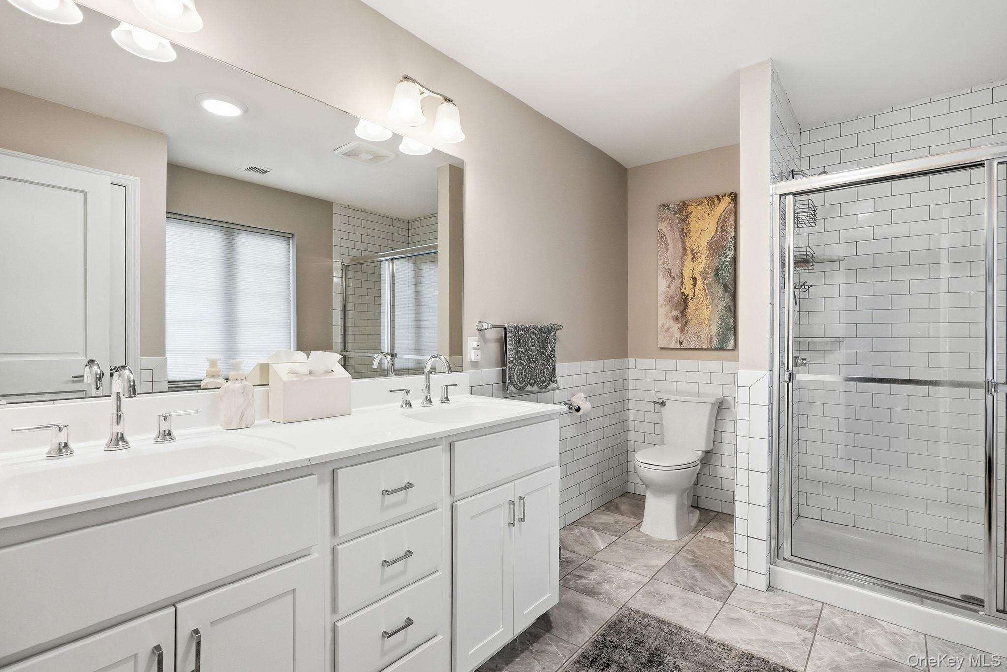 8 Muscoot River Road Granite Springs, NY 10527 - Photo 22 of 40 Primary bathroom featuring a double vanity with white cabinetry and chrome fixtures, subway tile wainscoting, a glass-enclosed shower with subway tile surround, and large format floor tiling