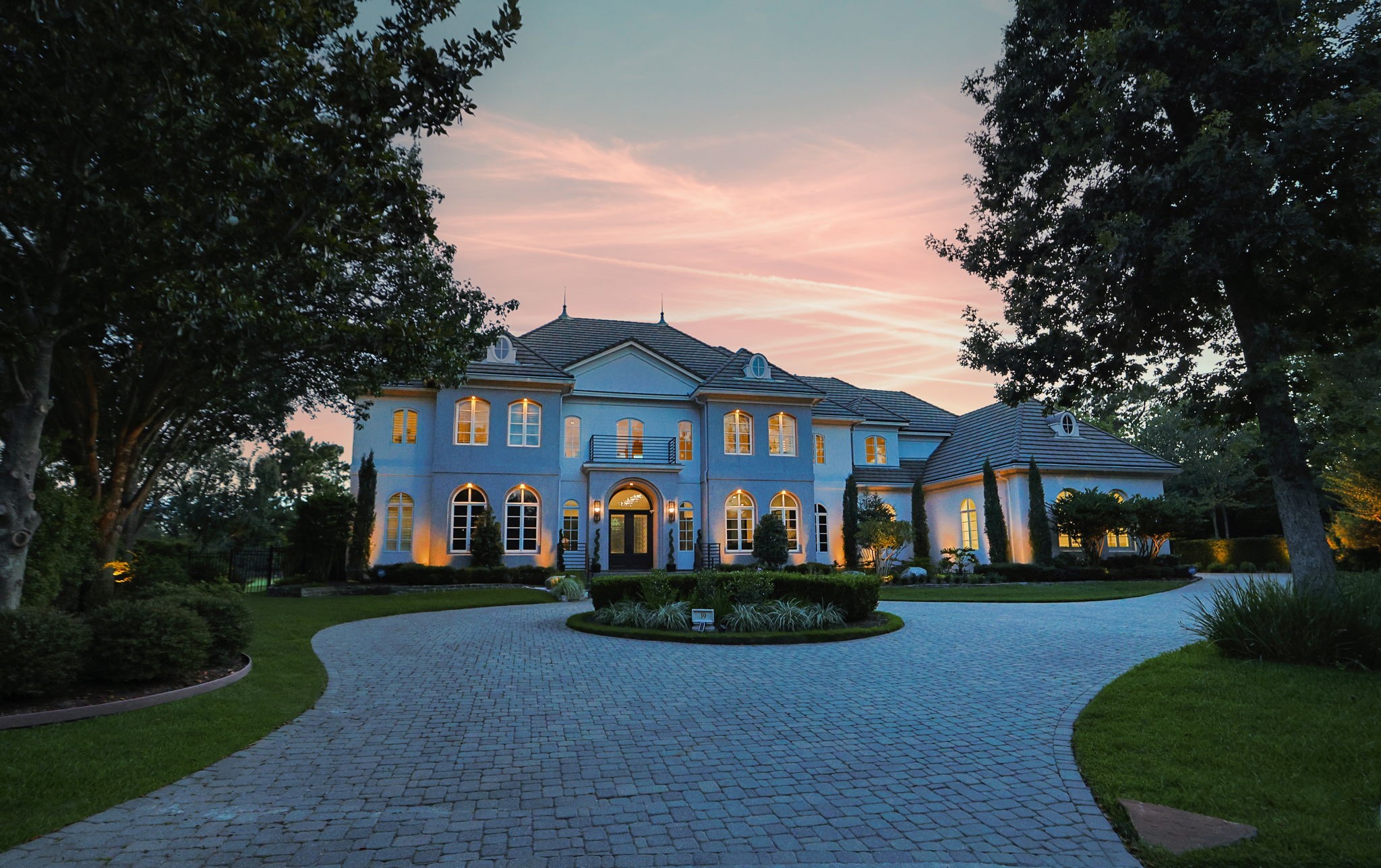 39 Maymont Way Spring, TX 77382 - Photo 1 of 50 This elegant two-story home features a grand facade with large windows and a stately entrance. It's surrounded by lush landscaping and a curved driveway, all under a picturesque sunset sky.