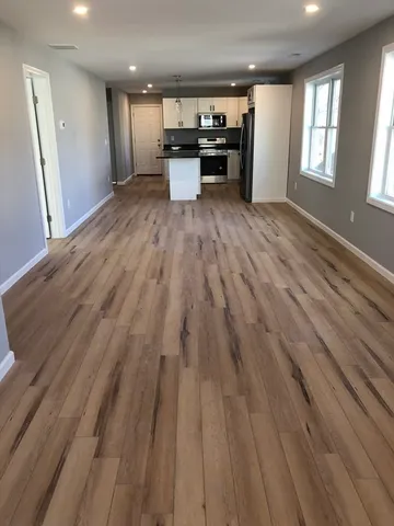 an empty room with wooden floor kitchen view and windows