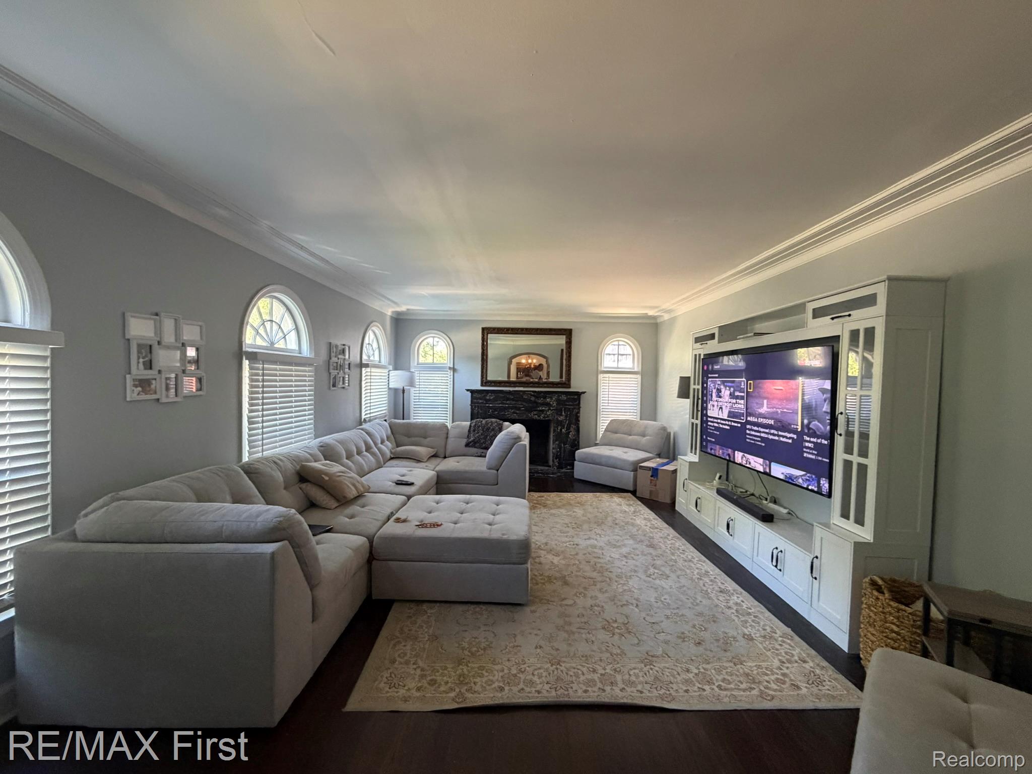 1349 Devonshire Road Grosse Pointe, MI 48230 - Photo 5 of 26 Spacious living area featuring dark wood-finish flooring, multiple arched windows with blinds, crown molding, and a built-in media console with glass-front cabinetry
