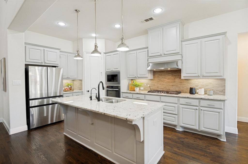 14215 Raspberry Lane Frisco, TX 75035 - Photo 6 of 30 Gourmet kitchen featuring a central island with a light-toned countertop, dark wood-finish flooring, white cabinetry, integrated stainless steel appliances, and a textured tile backsplash