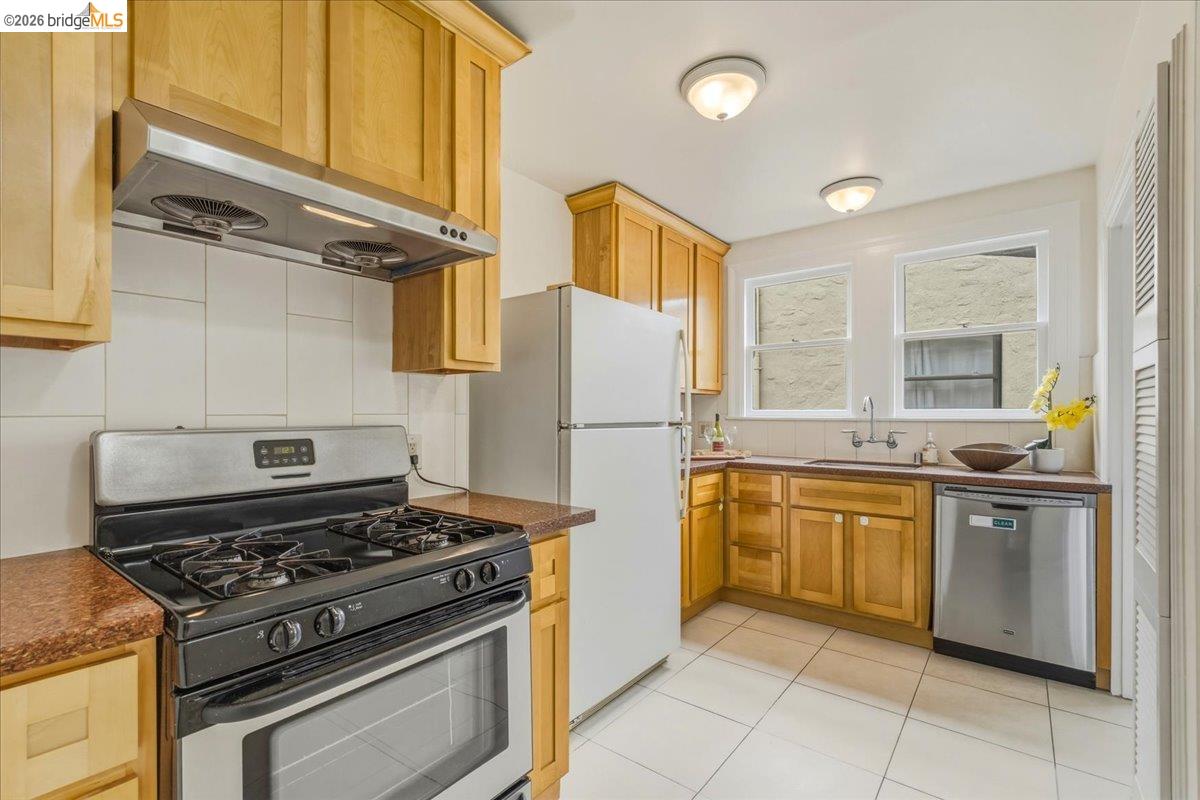 2403 Cedar Street Berkeley, CA 94708 - Photo 9 of 33 Kitchen featuring natural wood cabinetry, white tile flooring, dual windows, stainless steel dishwasher, and a built-in range with an overhead exhaust hood