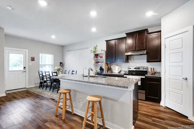 a kitchen with stainless steel appliances granite countertop wooden cabinets a refrigerator a sink a stove and chairs with wooden floor