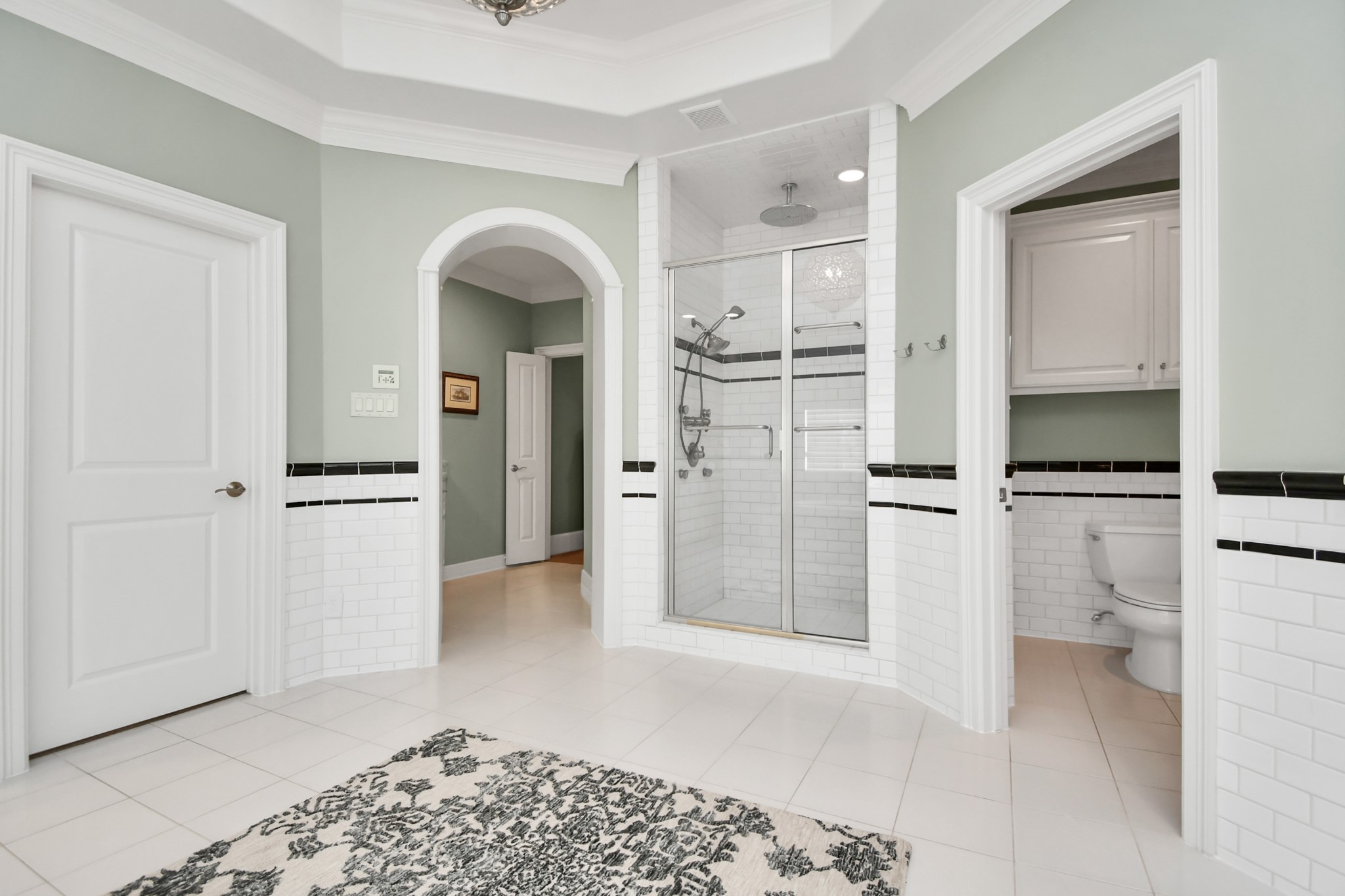 3739 Westerman Street Houston, TX 77005 - Photo 26 of 44 This elegantly designed bathroom features a spacious layout with a stylish walk-in shower, complete with rain shower and body jets. The shower area is adorned with classic white subway tiles accented by a chic black trim, adding a touch of sophistication. Adjacent to the shower, the bathroom includes a separate toilet area for added privacy. The room is painted in a soothing sage green, complemented by white wainscoting and black trim. A large arched doorway leads to an adjoining coffee bar.