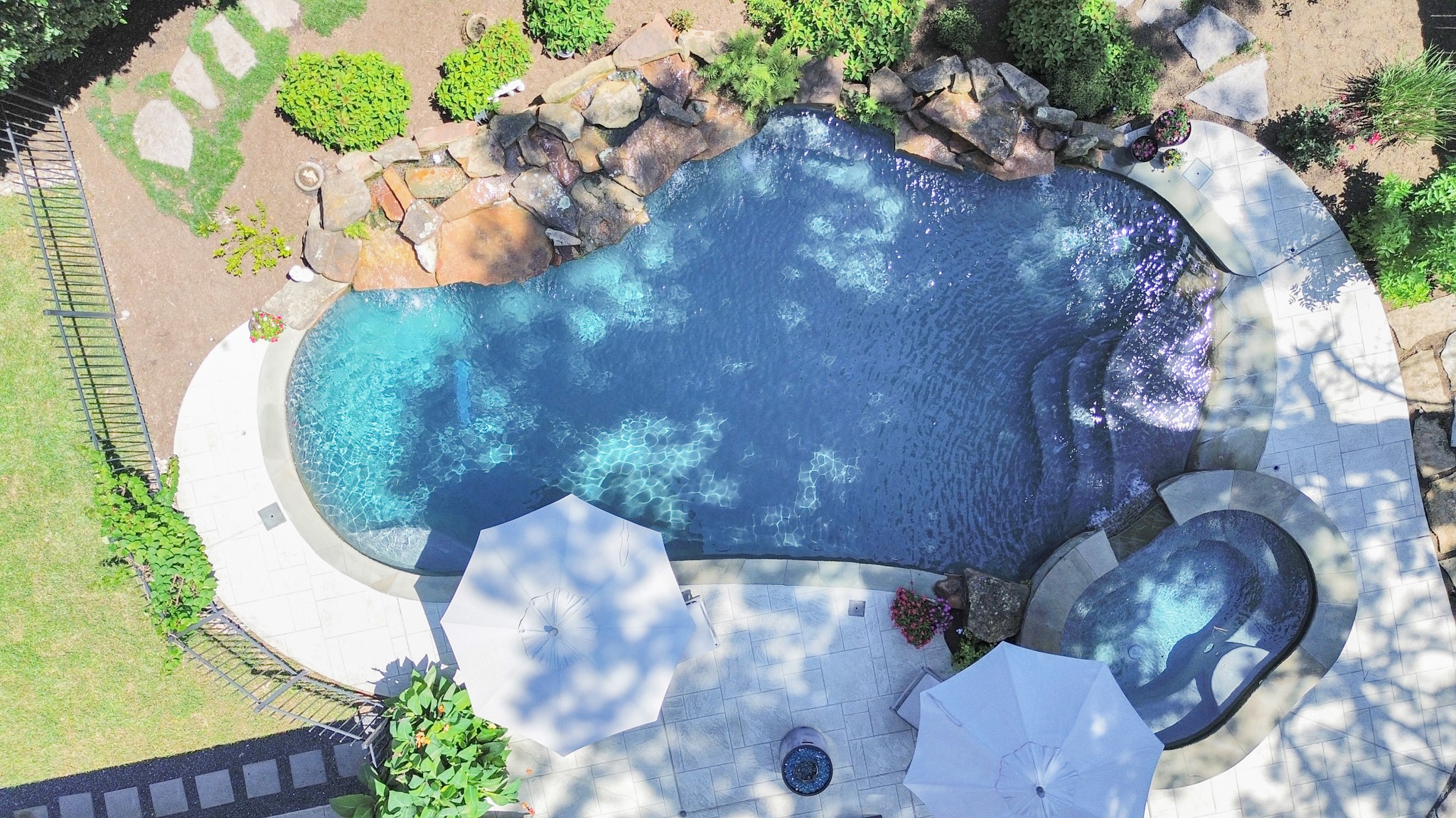 3739 Westerman Street Houston, TX 77005 - Photo 40 of 44 Aerial view of a luxurious backyard retreat featuring a sparkling rock pool with a cascading waterfall, surrounded by lush greenery and shaded by large umbrellas. The inviting hot tub adds a touch of relaxation to this stunning outdoor oasis.