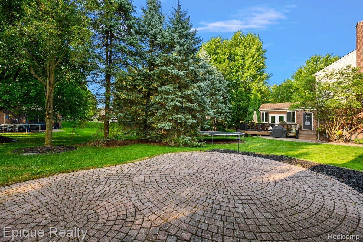 1909 Catlin Drive Rochester, MI 48306 - Photo 61 of 76 Circular paver patio surrounded by lush green lawn, mature trees, and a spacious outdoor deck with direct access to the interior
