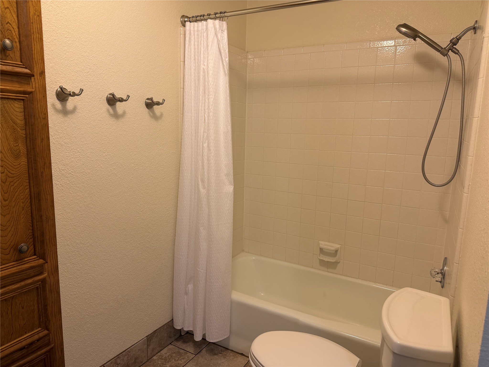 349 Fantail Loop, Unit B Austin, TX 78734 - Photo 7 of 9 Built-in wood cabinetry with panel detailing, a white ceramic tub and shower with tile surround, a wall-mounted showerhead with handheld sprayer, a ceramic toilet, and floor tiling