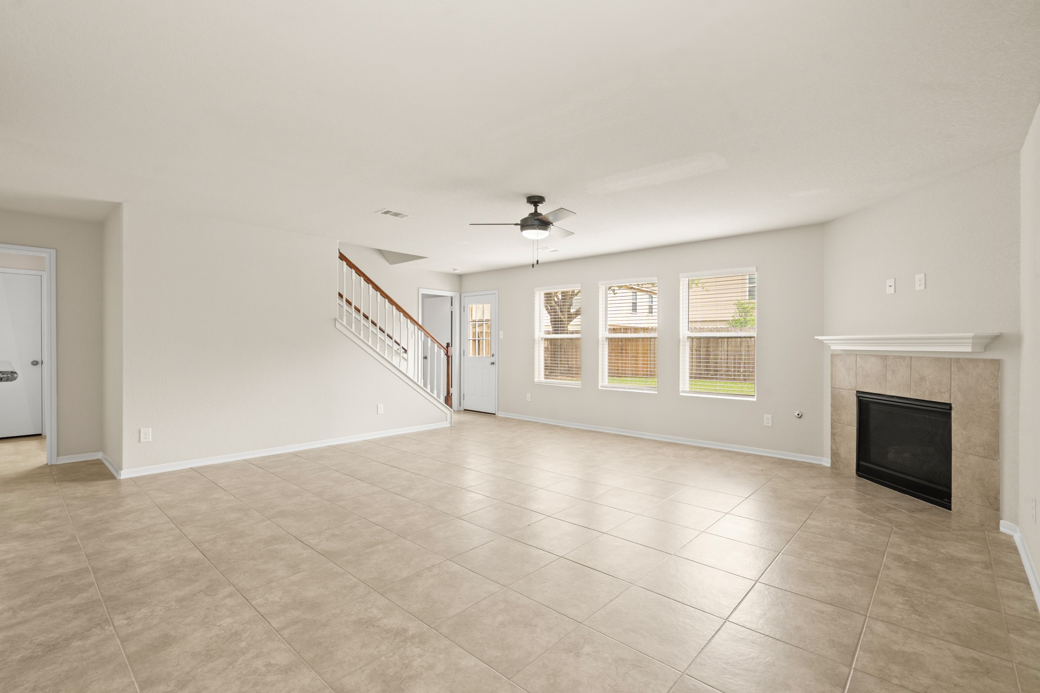 3118 Winchester Rnch Trail Katy, TX 77493 - Photo 4 of 22 The generously sized family room is the heart of this home, featuring a classic tile-surround fireplace with white mantel, a modern ceiling fan, gleaming tile floors, and a bank of windows that frame a lovely green backyard view. The staircase with white spindles and wood handrail adds architectural charm and character to the space.