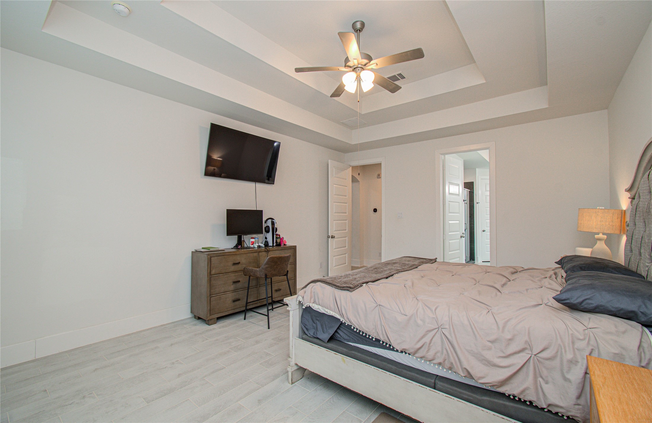4211 Limestone Terrace Manvel, TX 77578 - Photo 17 of 33 This spacious bedroom features a modern tray ceiling with a ceiling fan, light wood flooring, and a wall-mounted TV. It includes a comfortable bed, a dresser with a desk area, and an en-suite bathroom for convenience.