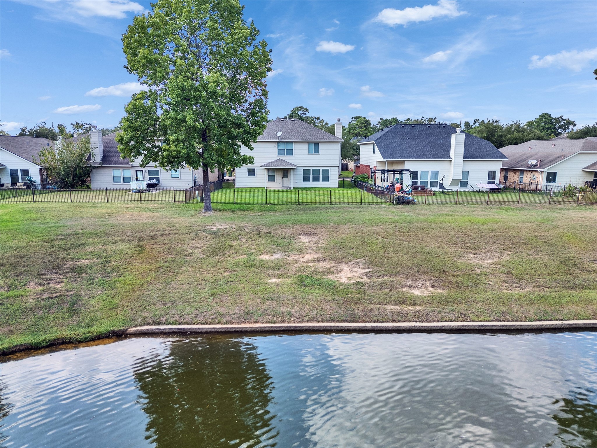 3307 Manor Tree Lane Houston, TX 77068 - Photo 37 of 44 This photo shows a two-story house with a fenced backyard facing a serene pond. The property features a large tree and is surrounded by similar homes, offering a peaceful suburban setting.
