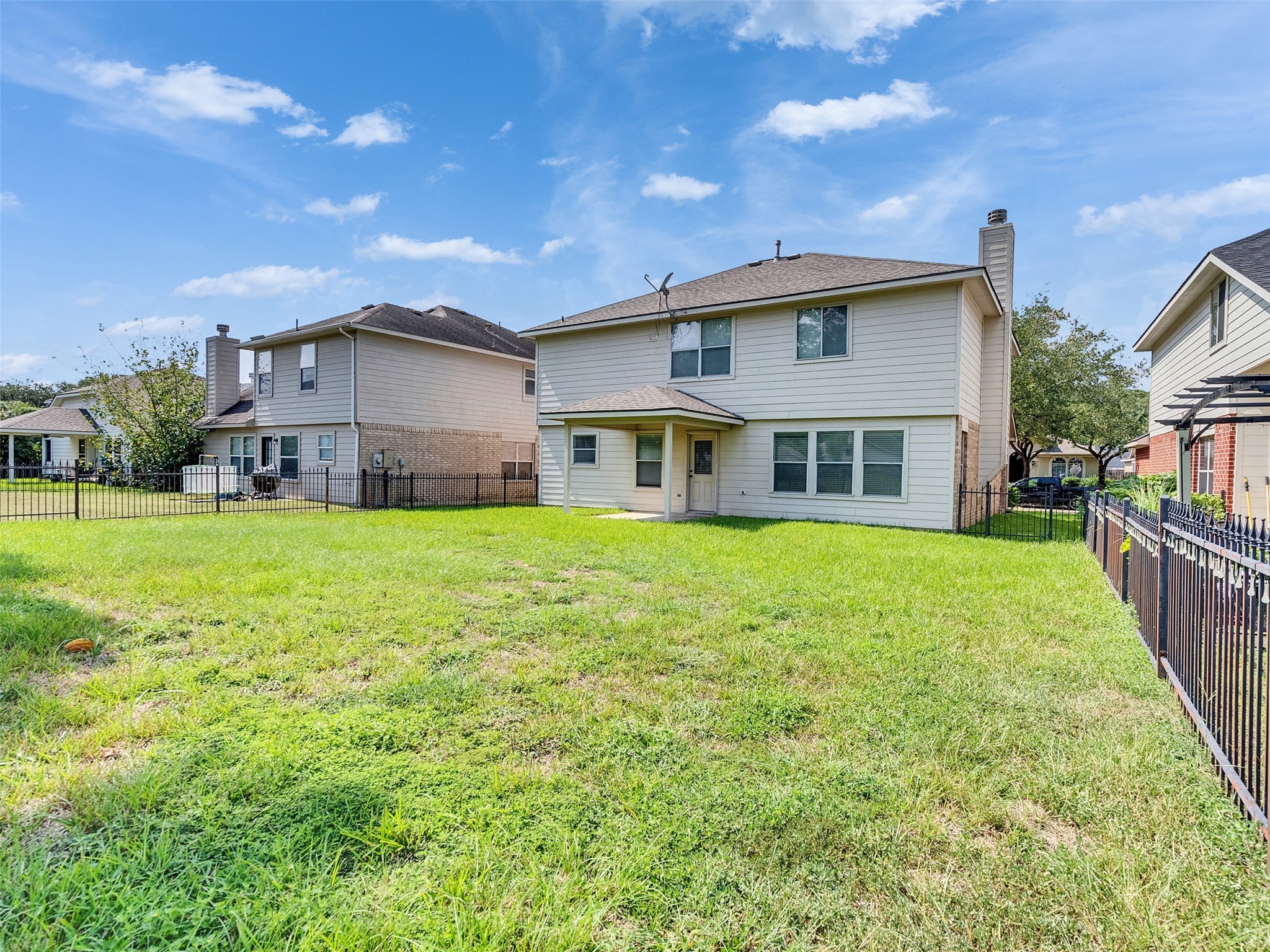 3307 Manor Tree Lane Houston, TX 77068 - Photo 39 of 44 The photo shows the backyard of a two-story home with a spacious, grassy lawn enclosed by a black metal fence. The house features multiple windows and a covered back porch, offering a private outdoor area perfect for family activities or entertaining.