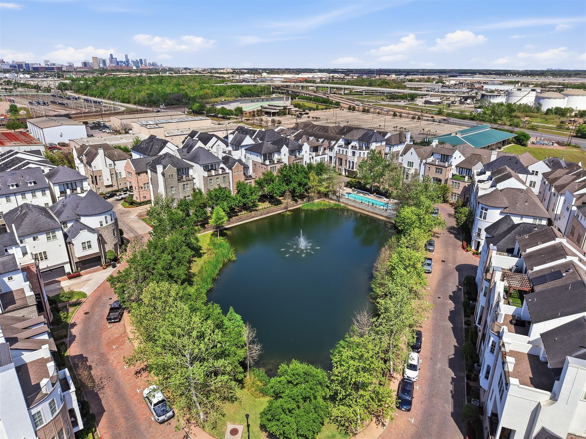 9427 London Bridge Station Houston, TX 77045 - Photo 30 of 30 This photo showcases a picturesque residential community with townhouses surrounding a serene pond featuring a central fountain. The area is lush with greenery and includes a small pool. The setting is urban, with city views in the background, providing a blend of tranquility and convenience.