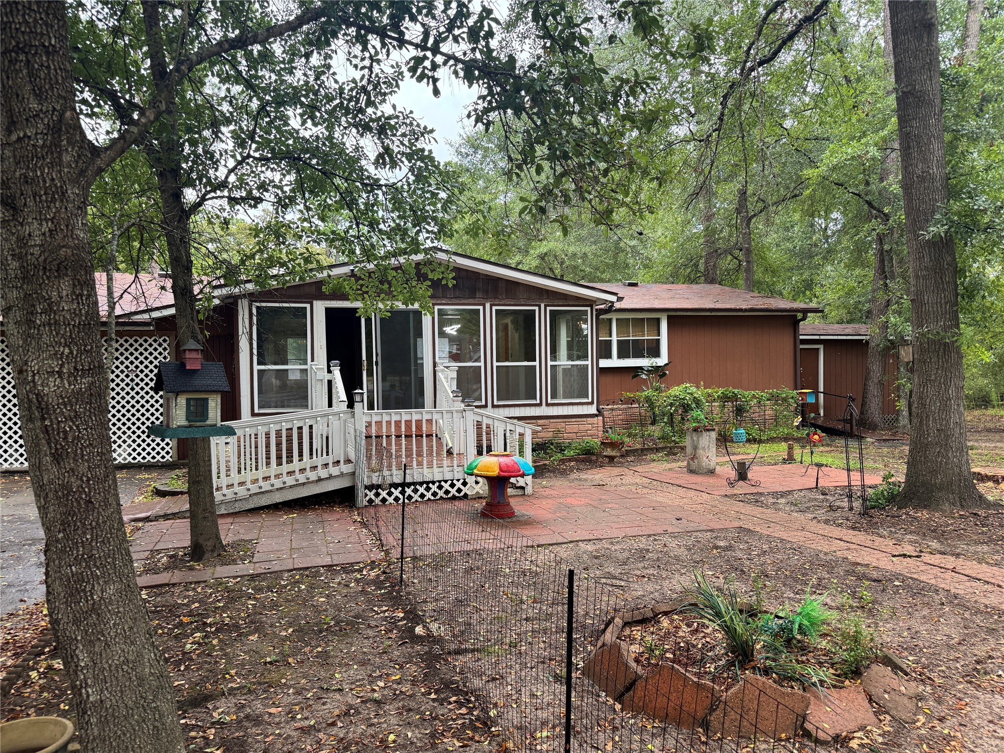 Charming rustic home surrounded by mature trees, featuring a cozy porch and a spacious patio area for outdoor enjoyment. The yard is partially fenced with garden space, creating a serene natural setting.
