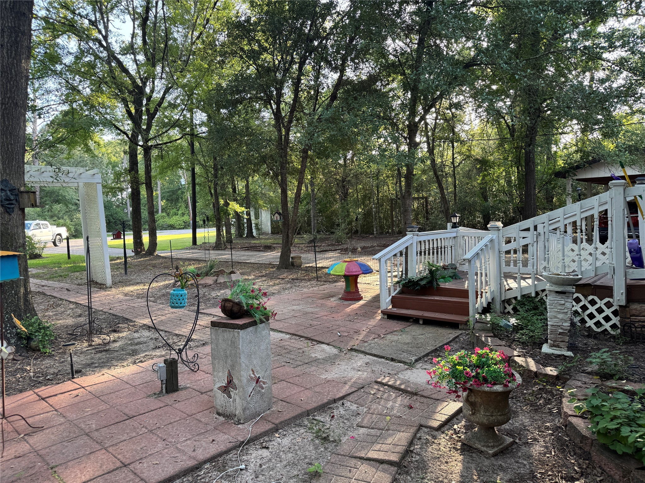 106 Westlake Road Trinity, TX 75862 - Photo 3 of 36 Charming backyard with mature trees, a decorative pathway, and a white railing leading to a small deck. Features include potted plants, garden decor, and a peaceful, wooded setting, offering privacy and a cozy outdoor space.
