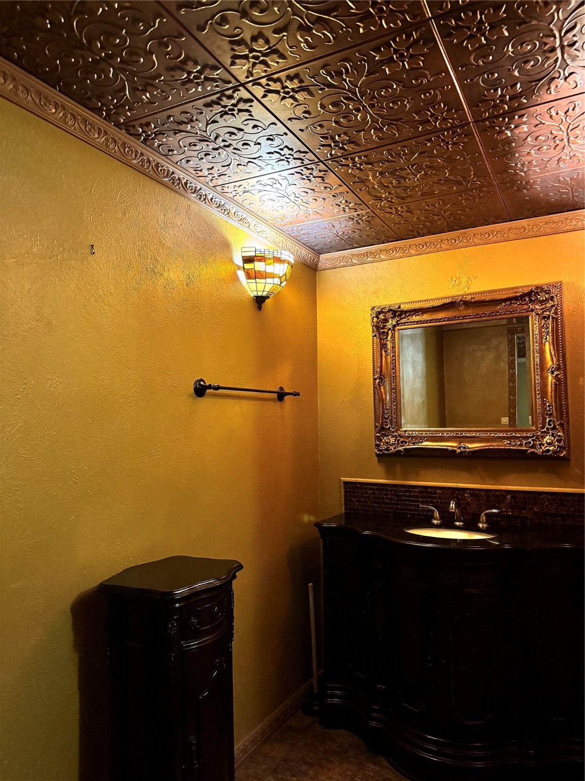 106 Westlake Road Trinity, TX 75862 - Photo 10 of 36 This bathroom features ornate metallic ceiling tiles, warm yellow walls, and a classic wooden vanity with an elegant framed mirror. A decorative wall sconce adds charm and soft lighting.