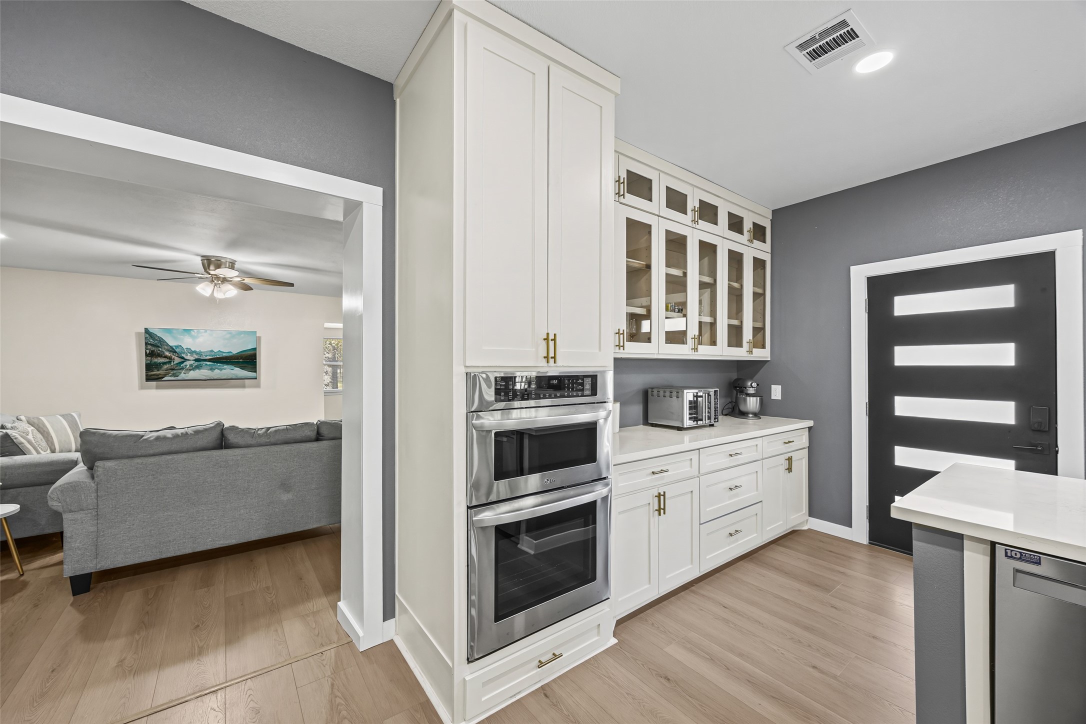 15080 Usher Lane Splendora, TX 77372 - Photo 18 of 44 This photo showcases a modern kitchen with sleek white cabinetry, stainless steel appliances, and a stylish glass-front cabinet. The space is complemented by light wood flooring and a contemporary dark door. A cozy living area with a gray sofa and ceiling fan is visible through an open doorway.