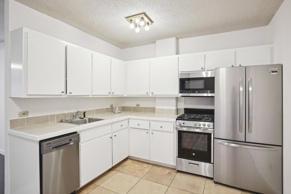 1212 Guadalupe Street, Unit 210 Austin, TX 78701 - Photo 2 of 12 Kitchen featuring appliances with stainless steel finishes, white cabinetry, tile countertops, a textured ceiling, and light tile patterned flooring