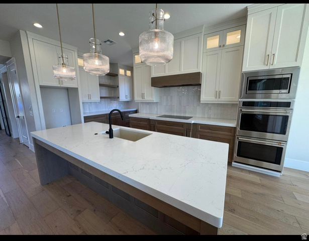 2788 East Rill Drive Washington, UT 84780 - Photo 6 of 41 Spacious kitchen featuring a large island with a composite sink and a matte black faucet, integrated stainless steel double ovens, a built-in microwave, wood-finish flooring, and white upper cabinetry with varied wood-finish lower cabinetry