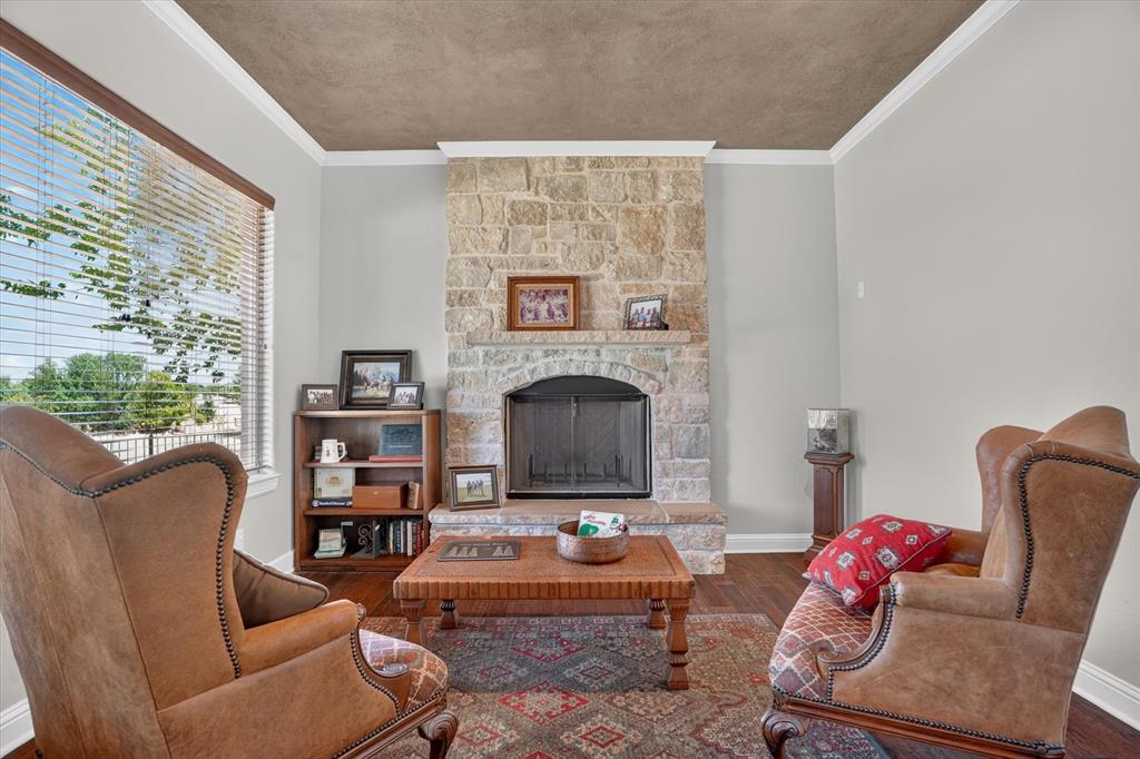 100 Canyon Ridge Trail Aledo, TX 76008 - Photo 21 of 38 Elegant living room showcasing a stone fireplace with dark metal surround and mantel display, paired wingback chairs with cognac leather upholstery, hardwood-finish flooring, geometric area rug, wooden coffee table, built-in shelving unit with display space, and coffered ceiling detail with neutral wall finishes