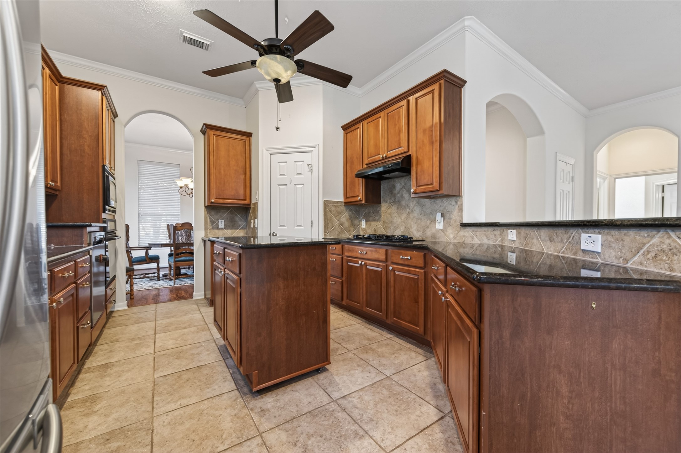 27 Sovereign Way Conroe, TX 77384 - Photo 14 of 49 This kitchen features rich wooden cabinetry, granite countertops, and a central island. It includes a tile backsplash, modern appliances, and a ceiling fan for comfort. Arched doorways connect to the dining area, enhancing flow and elegance.