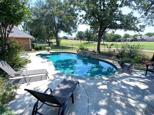 1762 Timber Ridge Circle Corinth, TX 76210 - Photo 26 of 26 Curved swimming pool with an integrated waterfall feature, surrounded by concrete decking and mature landscaping, with a black metal fence bordering a manicured green space