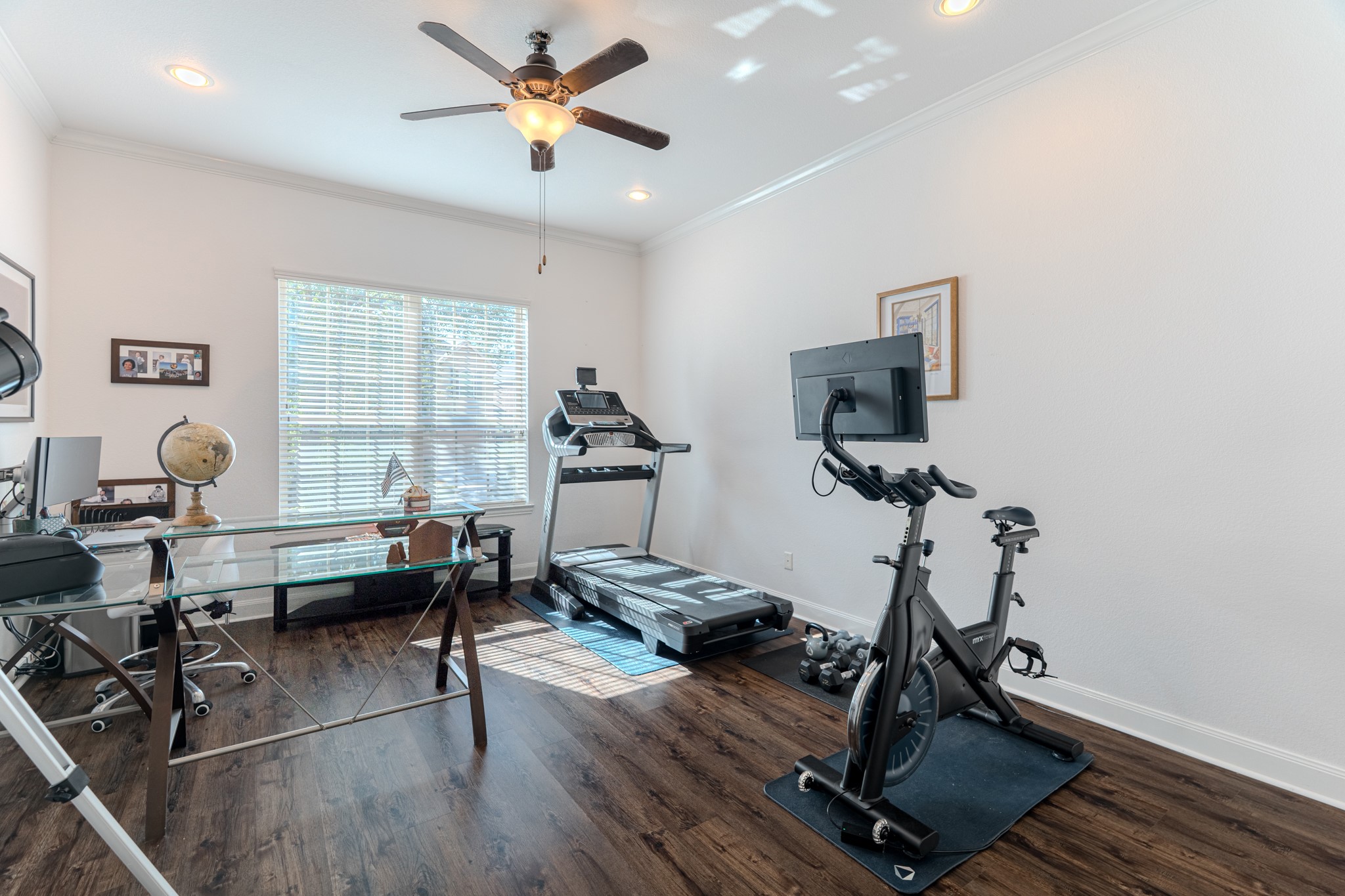 30 Fury Ranch Place The Woodlands, TX 77375 - Photo 13 of 47 This bright, multi-purpose room features a home office setup with a glass desk and a globe, alongside exercise equipment including a treadmill and stationary bike. The space has hardwood floors, a ceiling fan, and ample natural light from a large window.