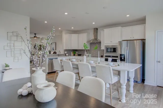 a large white kitchen with stainless steel appliances kitchen island granite countertop a dining table chairs and a refrigerator