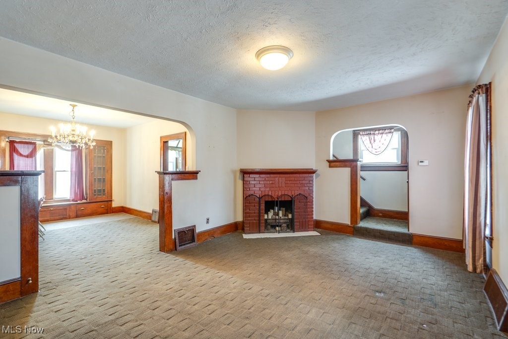 Unfurnished living room featuring a chandelier, carpet floors, a brick fireplace, stairway, and a textured ceiling
