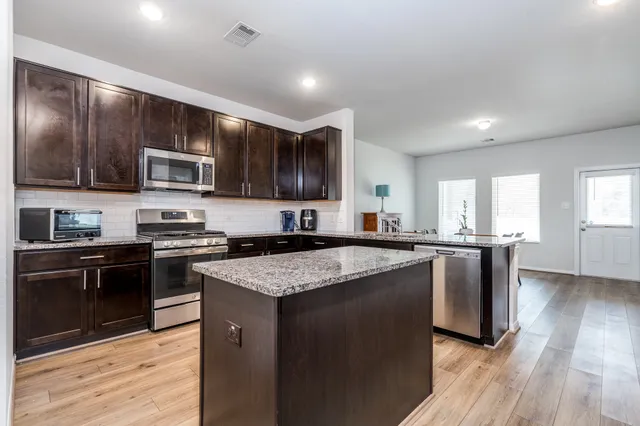 a kitchen with stainless steel appliances kitchen island granite countertop a dining table chairs and a refrigerator