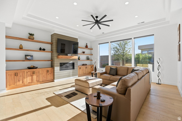 Living room featuring light wood-style flooring, a large fireplace, ceiling fan, recessed lighting, and a raised ceiling