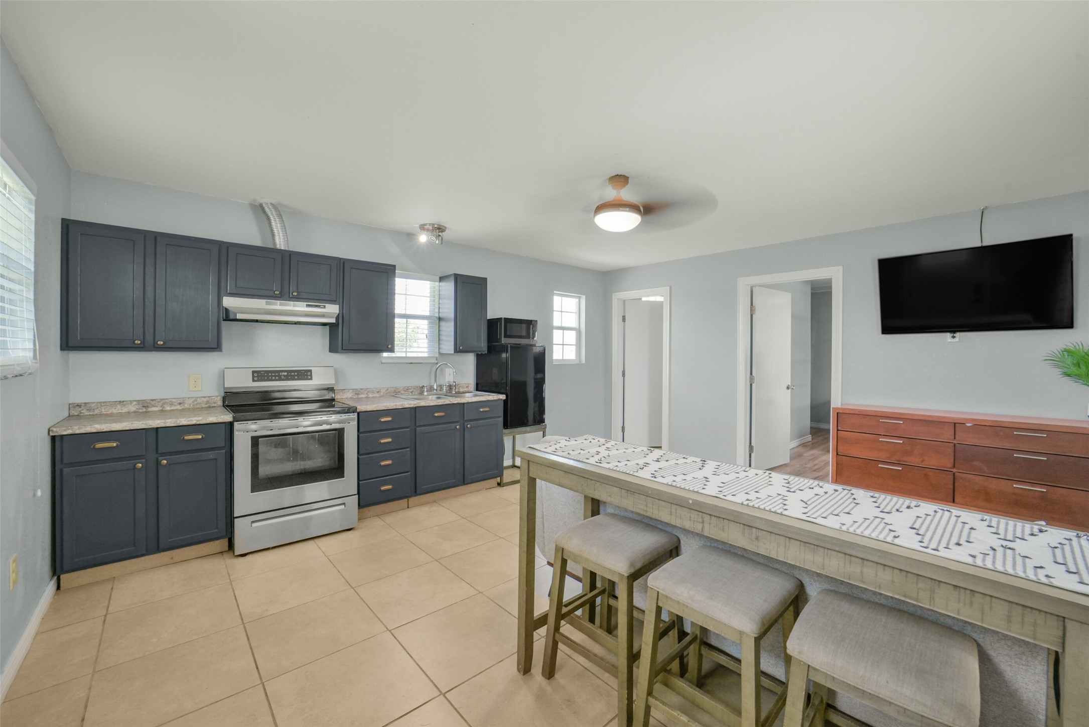 11301 South FM 441 Road El Campo, TX 77437 - Photo 27 of 43 Inside the Guest house, this kitchenette features modern appliances, dark cabinetry, and ample counter space. It includes a spacious breakfast bar with seating, tile flooring, and a ceiling fan for comfort. A wall-mounted TV and neutral walls create a welcoming and functional space.
