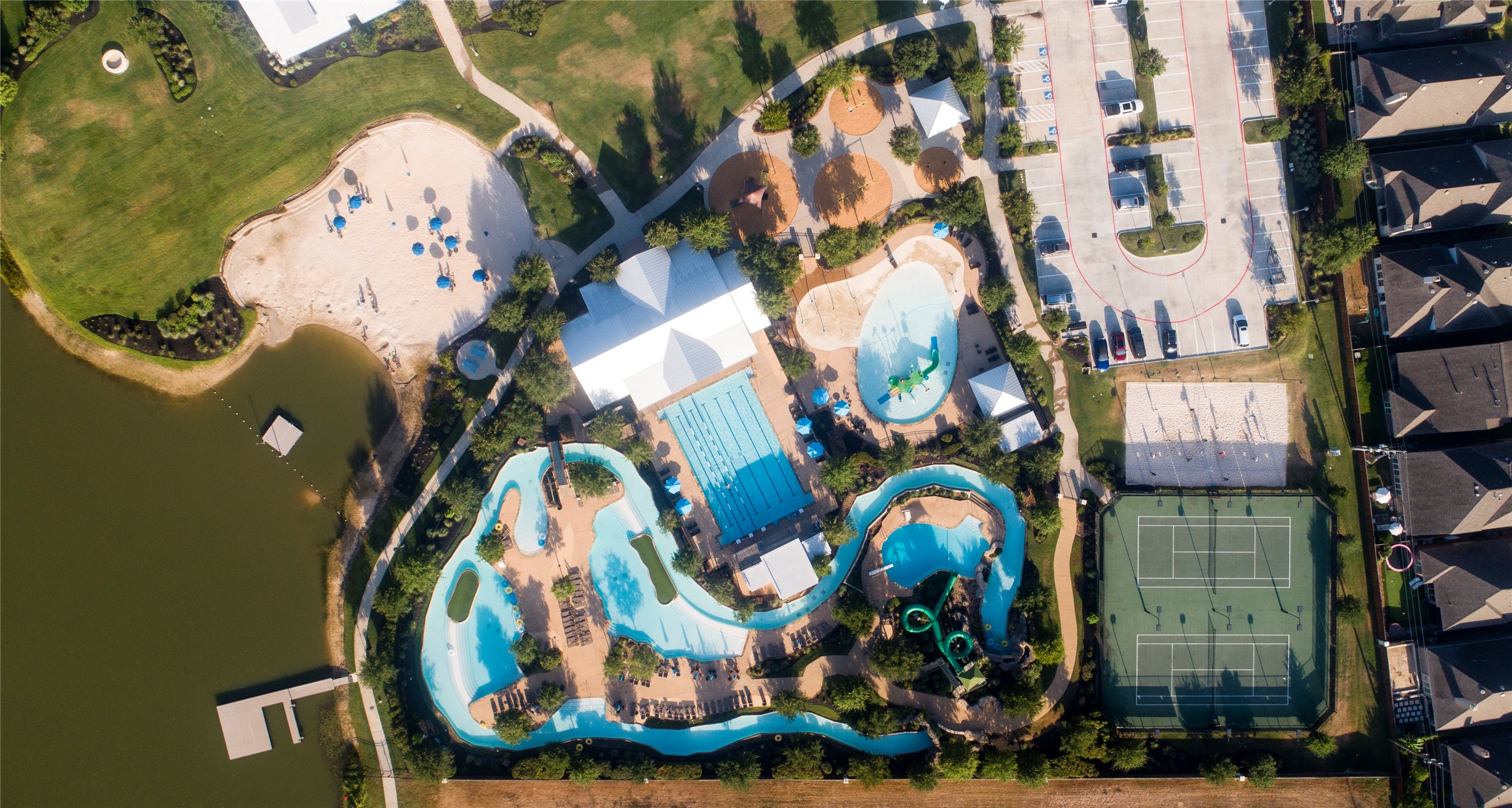 9438 Caddo Ridge Lane Cypress, TX 77433 - Photo 33 of 38 This aerial photo showcases a vibrant community recreational area featuring a large pool with a lazy river, water slides, a children's play area, tennis courts, and ample parking. It's adjacent to a lake and surrounded by lush greenery, offering a resort-like atmosphere for residents.