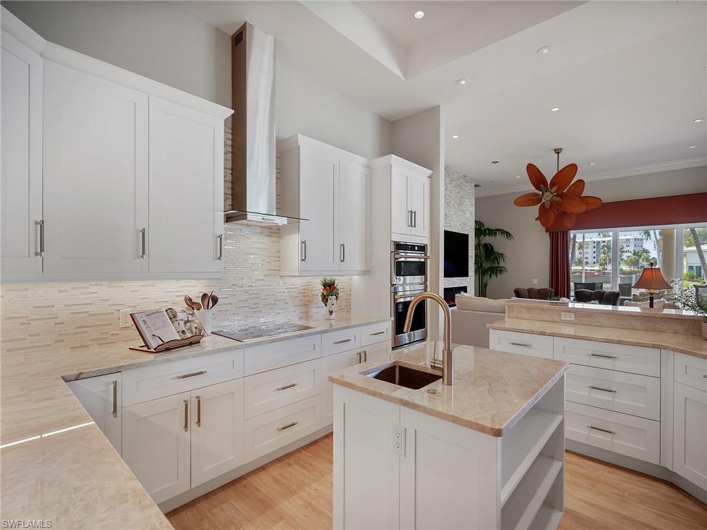 216 Springline Drive Naples, FL 34102 - Photo 6 of 46 Kitchen featuring open shelves, a center island with sink, decorative backsplash, wall chimney range hood, and light stone countertops