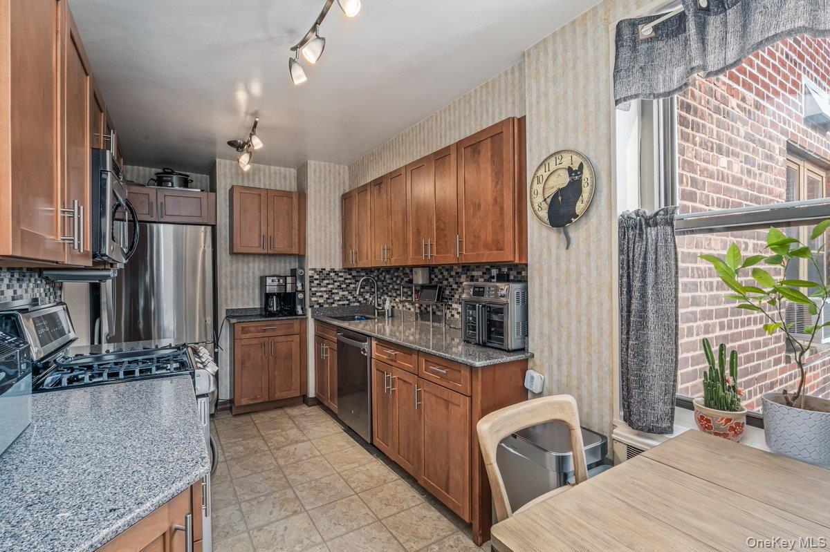 18-55 Corporal Kennedy Street, Unit 6H Queens, NY 11360 - Photo 4 of 18 Kitchen with stainless steel appliances, wallpapered walls, brown cabinetry, light stone counters, and tasteful backsplash