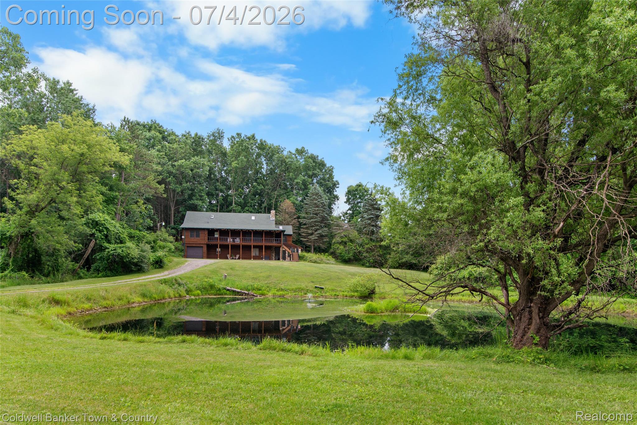 7335 Richardson Road Howell, MI 48843 - Photo 2 of 54 Expansive green lawn with a reflective pond, mature trees, and a two-story residence featuring wood siding, a wrap-around balcony, and an integrated garage
