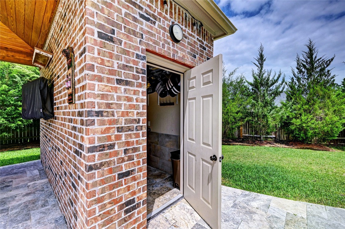 335 Fox Trail Road Montgomery, TX 77316 - Photo 30 of 47 This photo showcases a brick exterior with a partially open door leading to a half bath near the pool. It features a tiled travertine patio area and overlooks a lush, fenced backyard with trees, offering a sense of privacy and outdoor enjoyment.