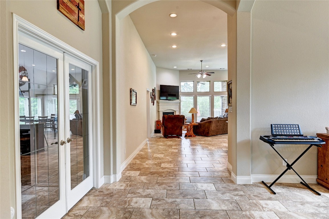335 Fox Trail Road Montgomery, TX 77316 - Photo 7 of 47 This photo showcases a spacious, well-lit hallway with tile flooring leading to a cozy living area. The living room features large windows, a ceiling fan, and a view into the kitchen through French doors on the left. The neutral color palette and open layout create an inviting atmosphere.