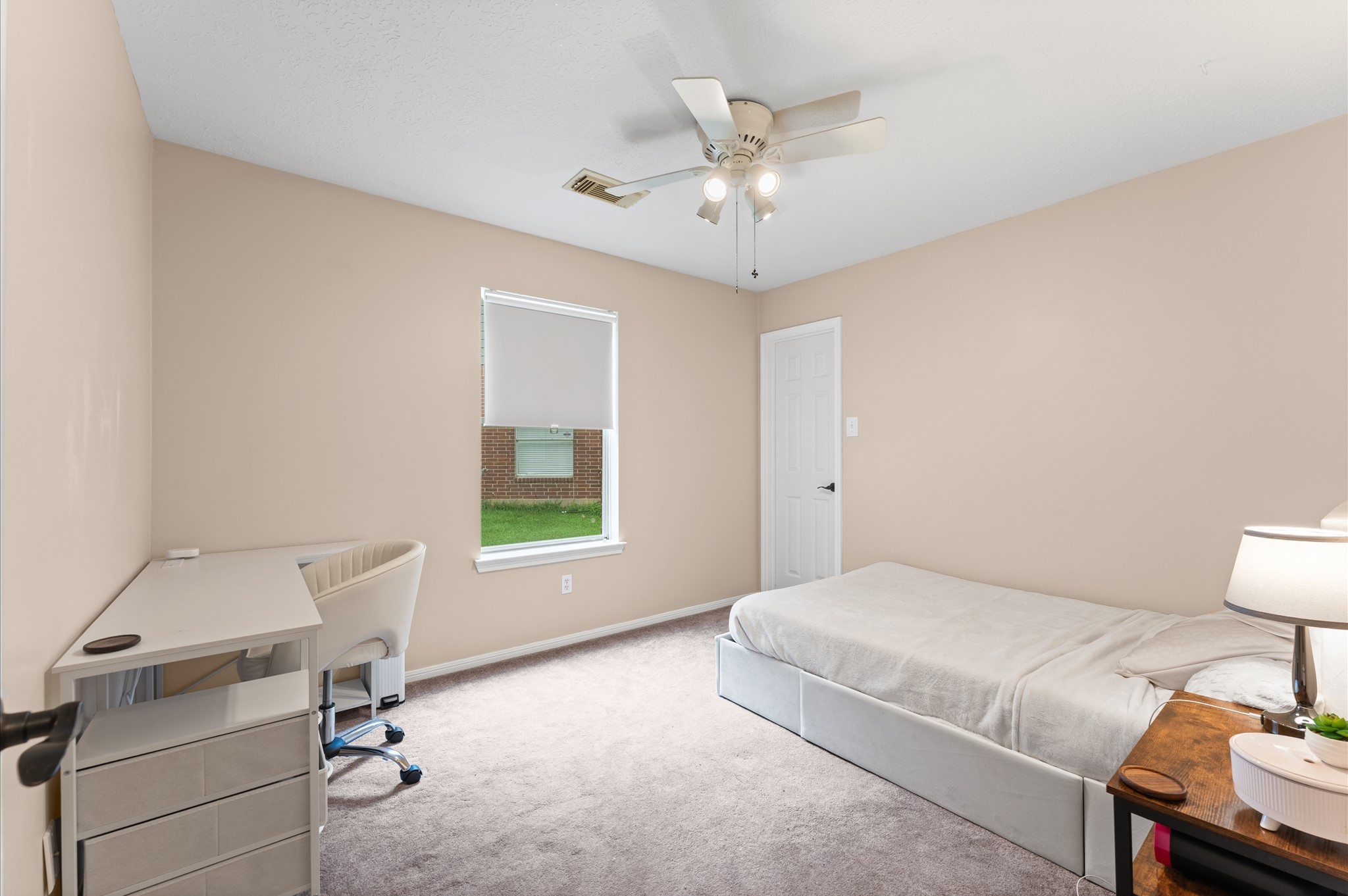 17618 Mill Springs Drive Spring, TX 77379 - Photo 17 of 26 This is a bright, neutral-colored bedroom featuring a ceiling fan, a window with a blind, a single bed, a small desk with a chair, and a bedside table with a lamp. The room offers a cozy and functional space.