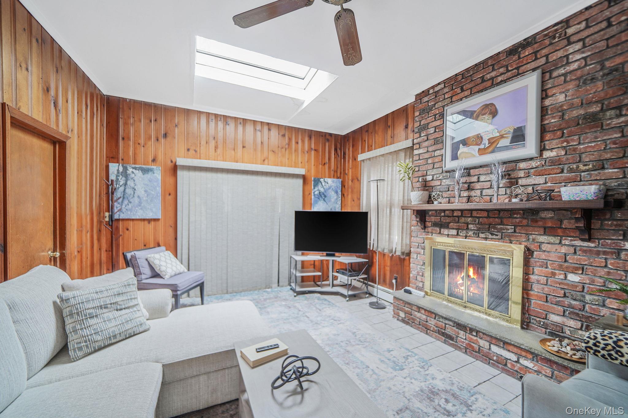 7 Rugby Road Westbury, NY 11590 - Photo 15 of 46 Living room with wooden walls, a brick fireplace, a skylight, tile patterned floors, and ceiling fan
