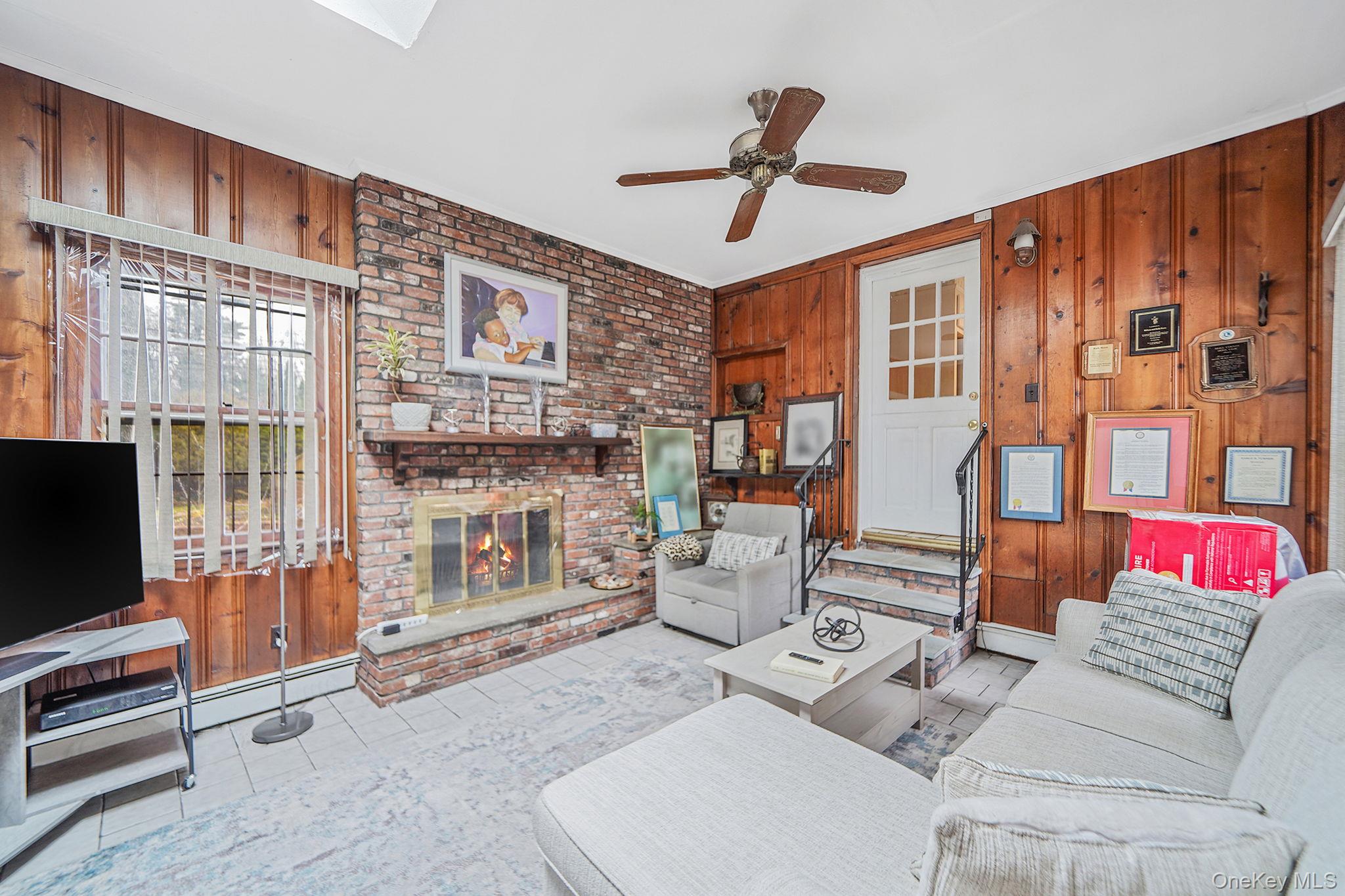 7 Rugby Road Westbury, NY 11590 - Photo 16 of 46 Living room featuring wood walls, a brick fireplace, a ceiling fan, tile patterned floors, and a baseboard heating unit