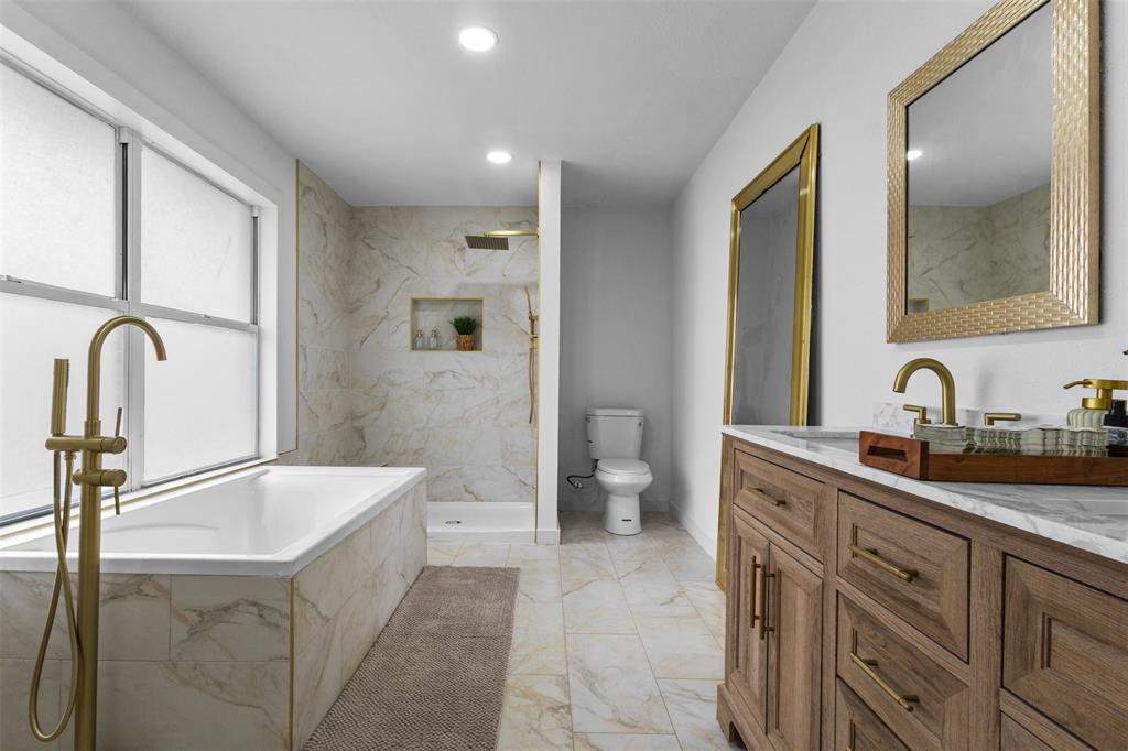 1917 South Vernon Avenue Dallas, TX 75224 - Photo 19 of 28 Spacious bath featuring a freestanding soaking tub with a floor-mounted brass faucet, walk-in shower with stone-look tile surround and recessed niche, dual vanity with brass fixtures and stone countertop, and large framed mirrors