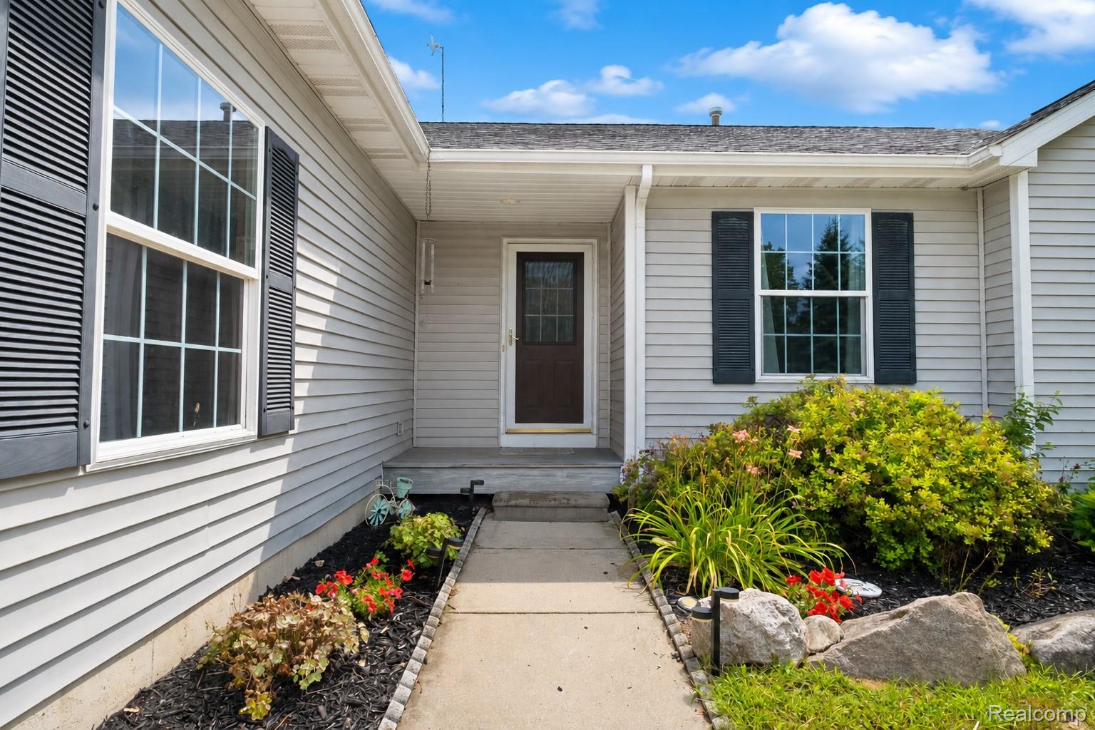 Light gray siding exterior with black louvered shutters, concrete walkway, and landscaped garden beds featuring various plantings