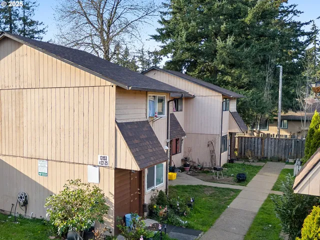 a view of a white house next to a yard with potted plants and big trees
