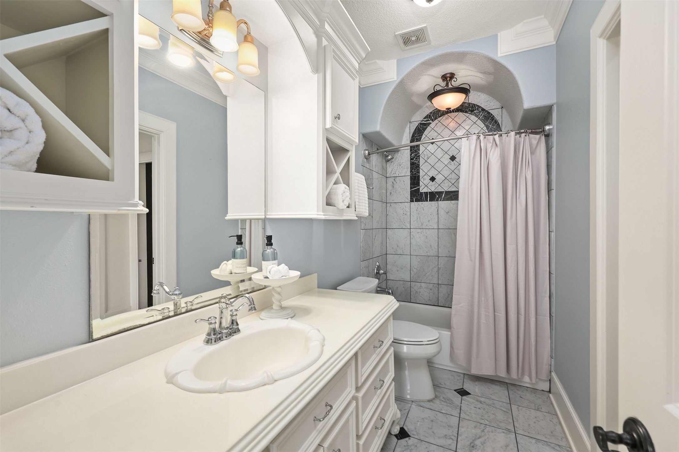 10896 Decatur Street Willis, TX 77318 - Photo 41 of 50 Discover a stylish bathroom oasis featuring light blue walls, elegant white cabinetry, and a large mirror, complemented by a tiled shower with an arched window, a classic vanity with modern fixtures, and soft, neutral accents—all beautifully illuminated for a clean and inviting atmosphere.