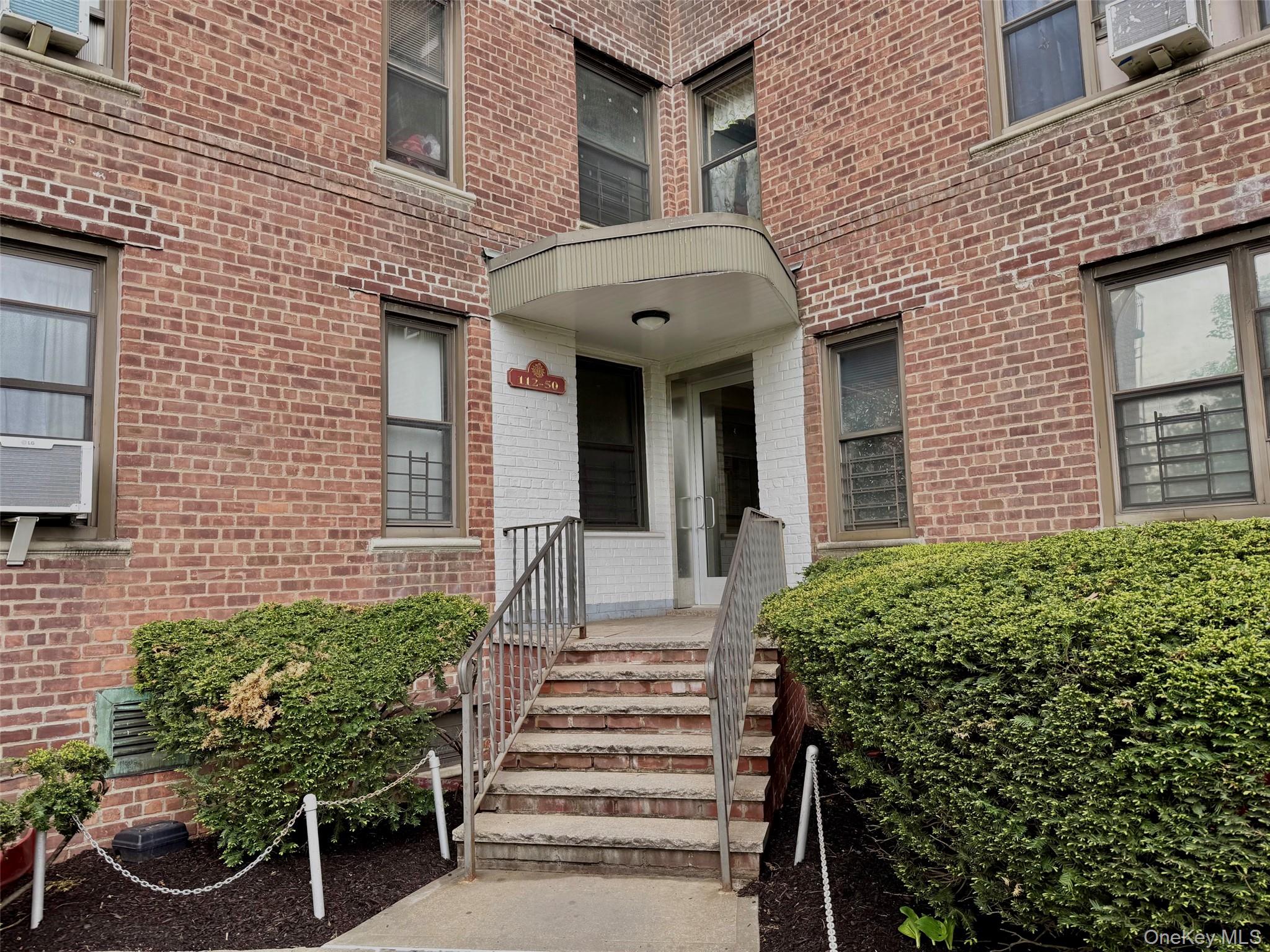 Brick facade building featuring a covered entrance, metal handrails, and established shrubbery