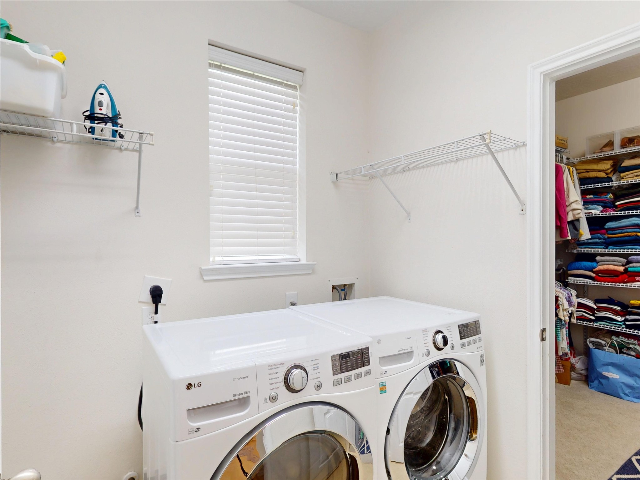 17402 Rainer Valley Lane Humble, TX 77346 - Photo 5 of 33 This photo shows a bright laundry room with modern front-loading washer and dryer units. It features a window with blinds, open shelving for storage, and an adjacent closet with neatly organized linens.