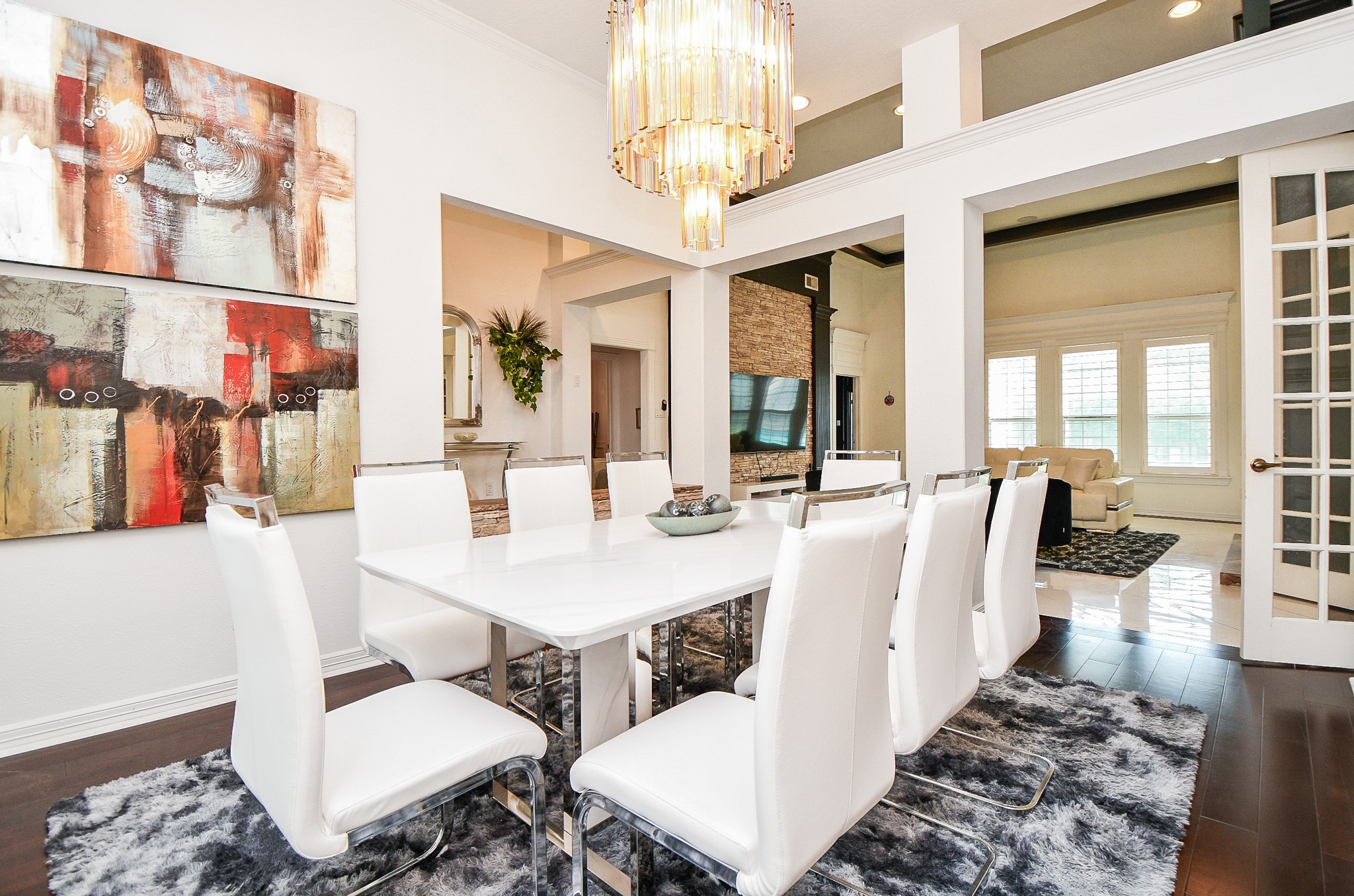 263 Wood Loop Street Houston, TX 77015 - Photo 22 of 45 This modern dining room boasts a sleek white table with eight matching chairs, set on a plush rug. It's accentuated by abstract artwork and a large chandelier. The space opens into a cozy living area with ample natural light from large windows and features elegant French doors.