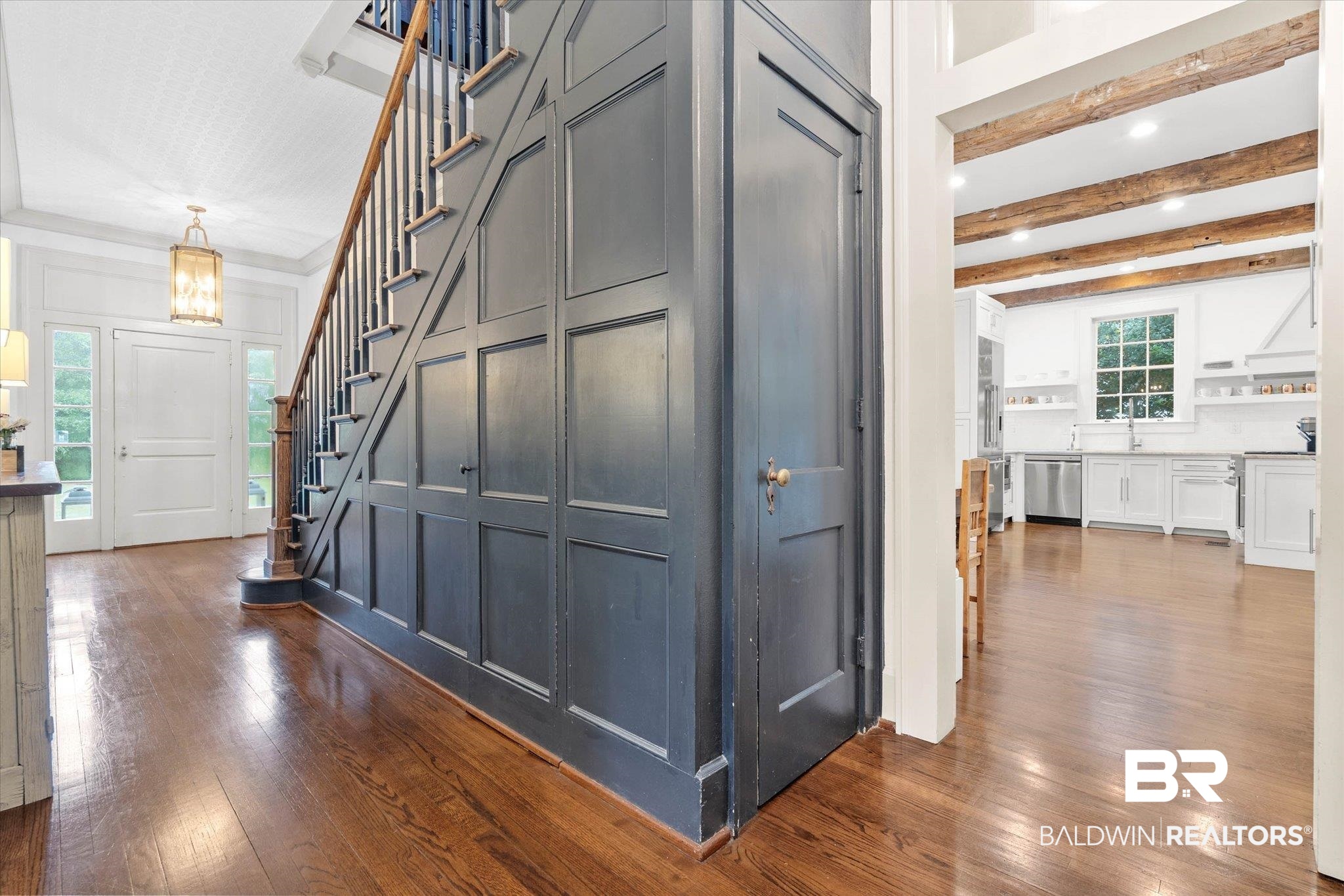 3863 Old Shell Road Mobile, AL 36608 - Photo 7 of 48 Hardwood flooring throughout the entry, staircase with wood paneling, and an adjacent kitchen featuring exposed beam ceilings, white cabinetry, and stainless steel appliances