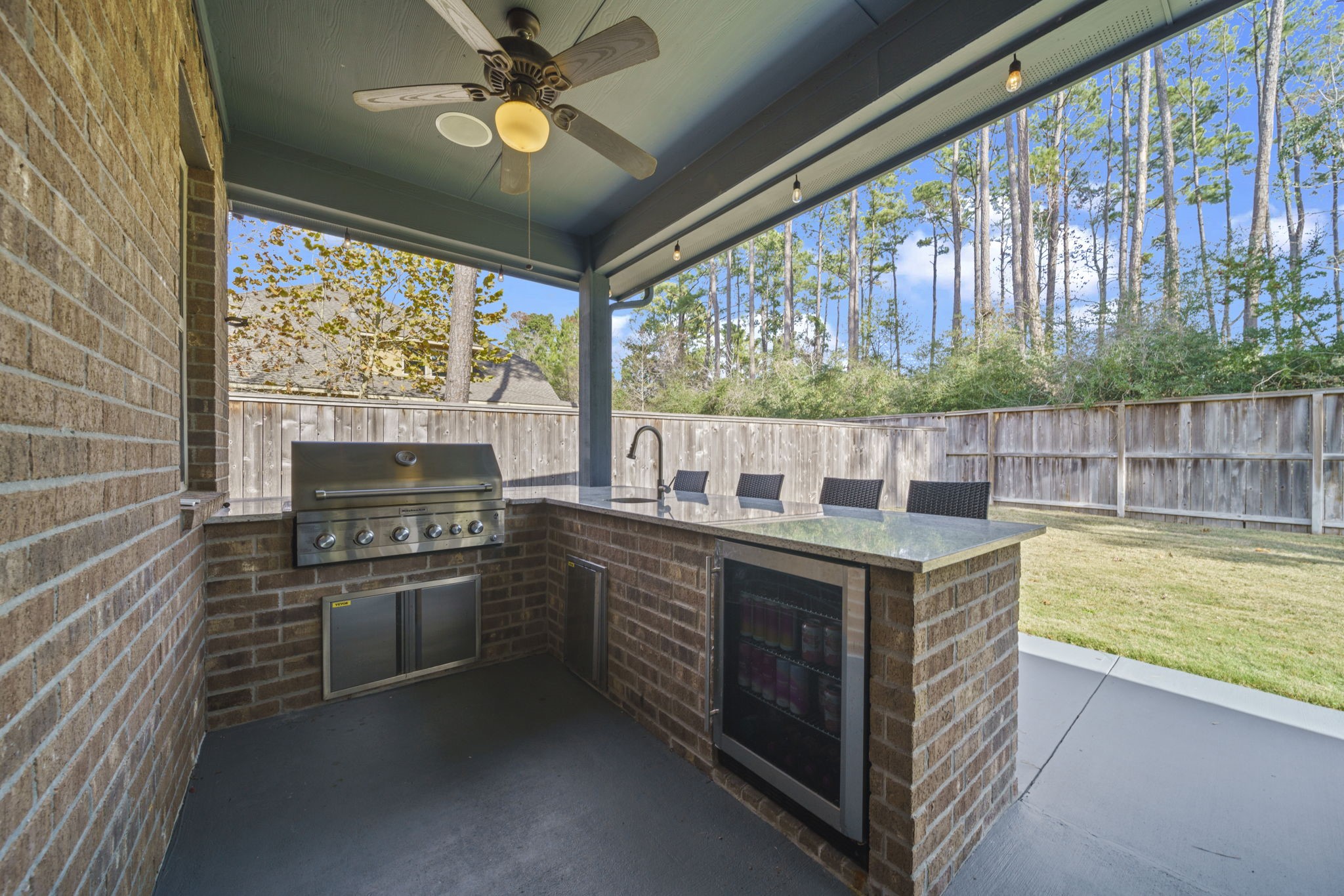 15 Inland Prairie Drive Tomball, TX 77375 - Photo 41 of 46 Enjoy this outdoor kitchen with a built-in grill, refrigerator, and bar seating under a covered patio. The space overlooks a fenced backyard surrounded by tall trees, perfect for entertaining and enjoying the outdoors.